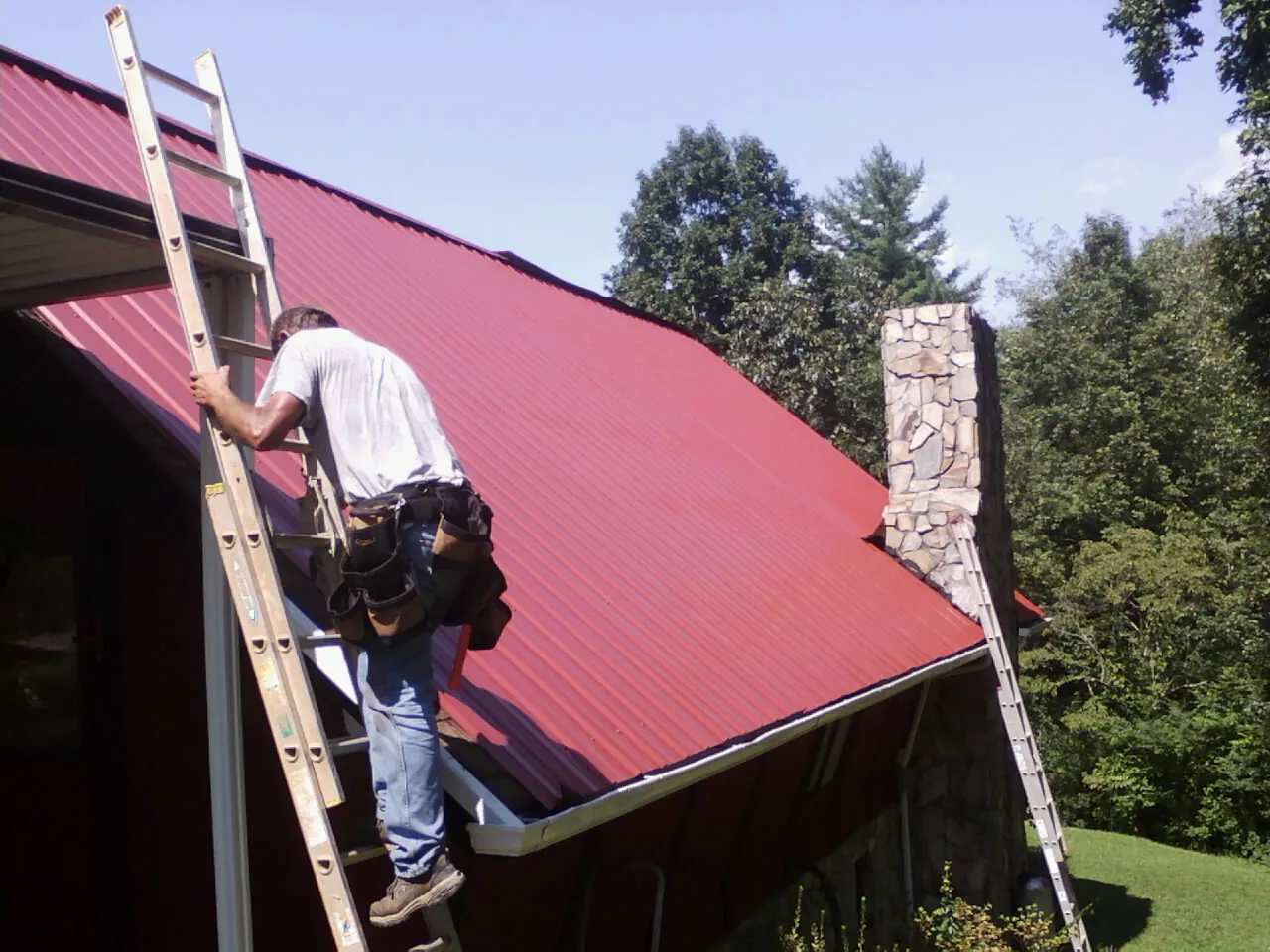 Roofer on a ladder beside a red metal roof and stone chimney — Soffit & Fascia Repair by Roofing Brothers in Renovo